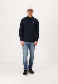 Cămașă navy cu mânecă lungă și butoni, având un logo galben pe piept, asortată cu blugi albaștri deschiși și adidași negri. Design simplu și ajustat.