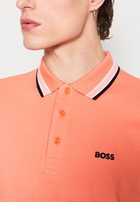 Korallenrotes Polohemd mit kurzen Ärmeln, aus strukturiertem Stoff, mit einem schwarz-weiß gestreiften Kragen und orangenen Knöpfen. "BOSS"-Logo in Schwarz.