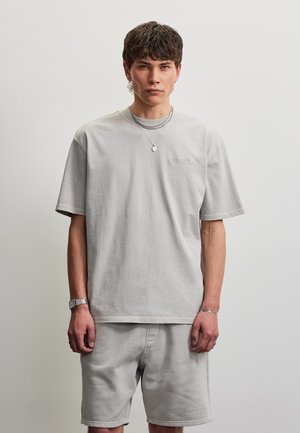 Jeune homme portant un t-shirt oversize gris clair et un short, accessoirisé avec un collier, une montre et un bracelet en argent, debout devant un mur blanc uni.