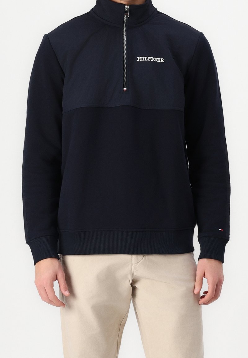 Homme portant un pull bleu marine à demi-fermeture éclair avec le logo "HILFIGER" et un pantalon beige, debout sur fond blanc.