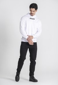 Mann in weißer Kapuzenjacke mit dem Text "GIANI KAVANAGH", schwarzen Hosen und schwarzen Sneakern, der vor einem schlichten hellgrauen Hintergrund steht.
