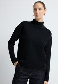 Kobieta z włosami związanymi w kok, w złotych kolczykach w kształcie kół, czarnym swetrze z wysokim kołnierzem i ciemnych dżinsach, trzymająca jedną rękę w kieszeni.