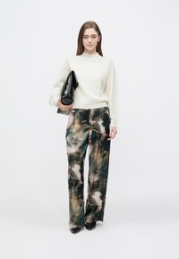 Modello che indossa un maglione crema con texture, pantaloni a gamba larga con motivo in verdi scuro e marroni, tiene in mano una pochette nera, con scarpe nere.