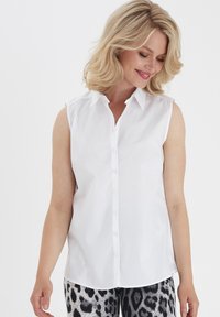 Cămașă albă fără mâneci, cu nasturi și guler, având o textură netedă și un design ajustat, asortată cu pantaloni cu un model alb-negru.