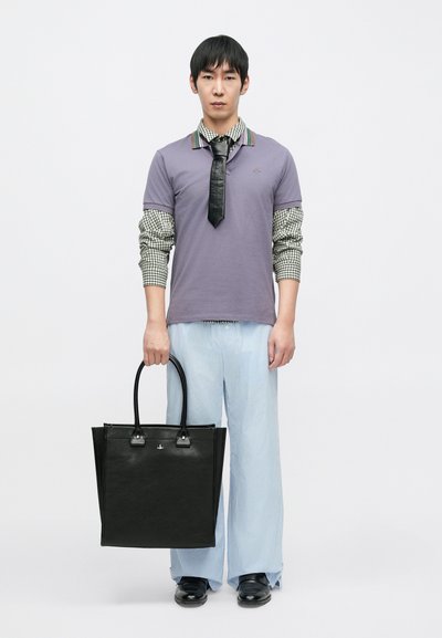 Fekete táska sima felülettel, ezüst színű fém részekkel és két fogóval. A modell lila galléros pólót és kék, csíkos, bő szárú nadrágot visel.