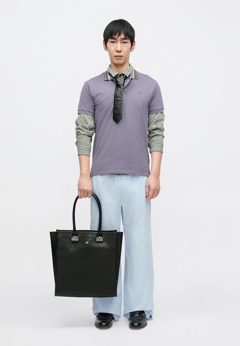 Black tote bag with a smooth texture, silver hardware, and two handles. Model wears a purple polo and striped blue wide-leg trousers.