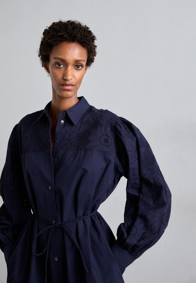 Granatowa sukienka koszulowa z zapięciem na guziki z przodu, z teksturowanym wyszywanym panelem i bufiastymi rękawami. Zawiera detal w postaci związanej talii.