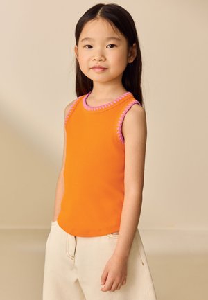 Junges Mädchen mit langen dunklen Haaren, das ein oranges ärmelloses Top mit pinkem Rand und helle Hosen trägt, steht vor einem beigen Hintergrund.