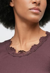 Γυναίκα με σγουρά μαλλιά που φοράει μια ριγωτή μωβ μπλούζα με κεντητό λαιμόκοψη.