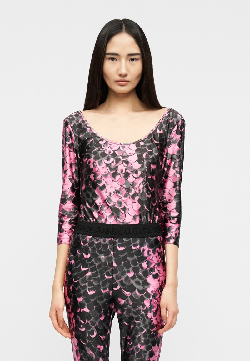 Γυναίκα με μακριά σκούρα μαλλιά που φοράει γυαλιστερό ροζ και μαύρο ρούχο με μοτίβο σαν λέπια και παντελόνι με ζώνη μέσης "Versace Jeans Couture".