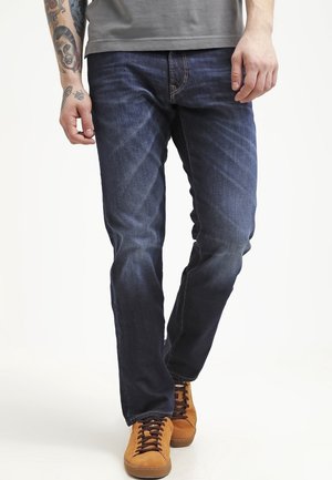 Mies, jolla on tummansiniset farkut, vaaleanruskeat mokkanahkaiset tennarit ja harmaa paita, näkyvillä tatuoitu vasen kyynärvarsi, kävelee valkoista taustaa vasten.