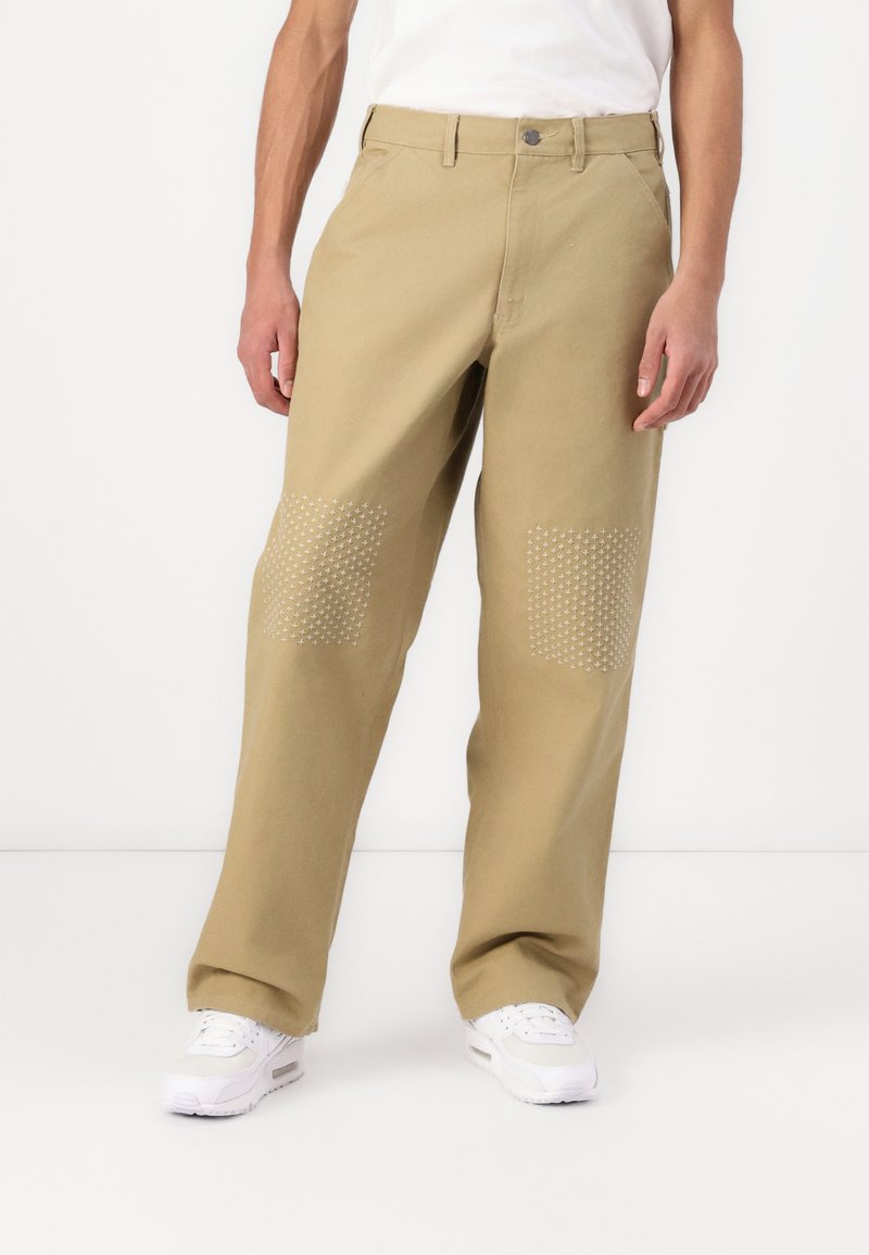 Beige bomullsbyxor med vida ben, prydda med vita stjärnmönstrade lappar på båda knäna. Designade med en klassisk midjeresår och framfickor.