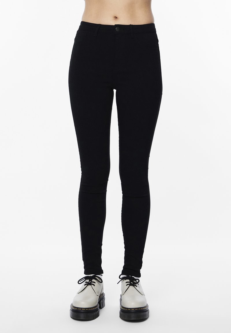 Pantalones ajustados negros con una textura de denim suave, cuentan con un cierre de botón y cinco bolsillos. Combinados con zapatos claros de cordones.