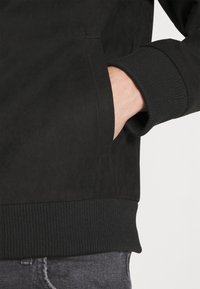 Black jacket in a soft, textured fabric with ribbed cuffs and waistband. Features a side pocket and a clean, minimalist design.