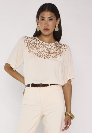Donna che indossa una camicetta crema con scollo floreale ritagliato, pantaloni crema, cintura marrone, orecchini vistosi e un braccialetto spesso, in piedi con la mano sull'anca.