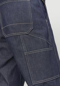 Tumedat sinilised denim püksid, millel on suured taskud, kontrastsed õmblused ning struktureeritud disain, kus on selgelt nähtavad õmblused ja kangatextuur.