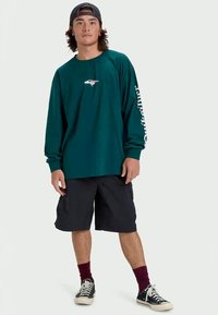 Long-sleeve teal shirt with a white logo on the chest, paired with black shorts. Model wears black sneakers and maroon socks.