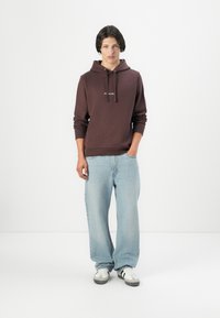 Brown cotton hoodie with kangaroo pocket and drawstring, paired with light blue denim jeans. Black and white sneakers complete the look.