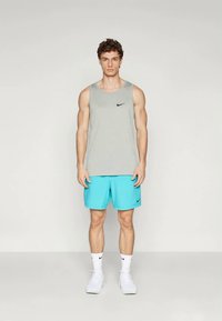 Fiatal férfi előre nézve áll, szürke ujjatlan Nike felsőt, türkiz Nike rövidnadrágot, fehér Nike zoknit és fehér sportcipőt visel.