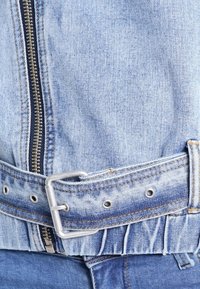 Světlemodrá džínová bunda s výrazným stříbrným zipem, kovovou přezkou a odpovídajícím páskem, s patrným šitím a mírně opotřebovanou strukturou.
