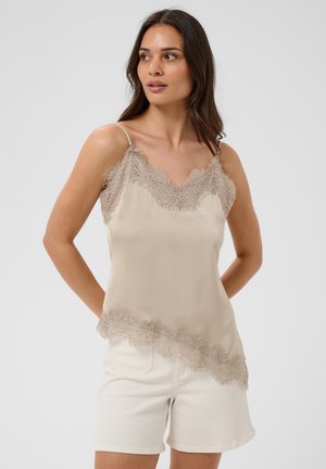 Donna che indossa una canottiera beige con bordo in pizzo e pantaloncini bianchi, in piedi con le mani dietro la schiena, guarda di lato.
