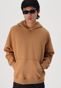Sudadera con capucha marrón, hecha de un tejido suave, con un bolsillo tipo canguro, puños y dobladillo acanalados. El modelo lleva gafas de sol negras.