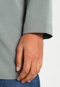 Maglietta a maniche lunghe di un grigio chiaro con una texture liscia e un polsino arrotolato. Una mano è posata sul polsino, rivelando una dettagliata texture della pelle.