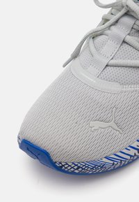 Light gray athletic shoe with a textured knit upper, reflective accents, and a blue patterned sole. Features logo on the side and laces.