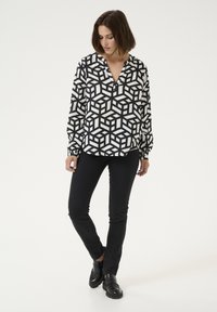 Zwart-witte blouse met een patroon van geometrische kubusontwerpen, lange mouwen, kraagloze halslijn, gecombineerd met zwarte slim-fit broek en schoenen.