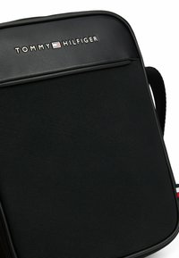Bolso negro de Tommy Hilfiger con una superficie texturizada, que presenta un logotipo de la marca prominente y acentos suaves a lo largo de los bordes.