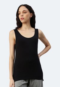 Canotta nera con scollo a pescatore, design senza maniche, texture liscia e orlo più lungo, abbinata a pantaloni grigi.