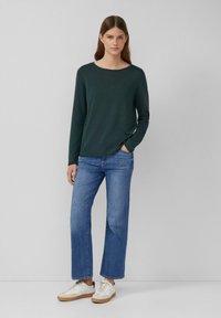 Dunkelgrüner Pullover mit langen Ärmeln, leichtes Strickmaterial, runder Ausschnitt; kombiniert mit blauen Schlagjeans und weißen Sneakers mit beigen Akzenten.