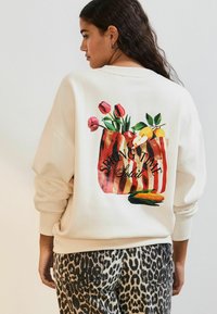 Huppari vaaleassa sävyssä, jossa on värikäs kukka- ja hedelmämotiivi selkäpuolella, sekä teksti "SPRING TIME Soleil." Yhdistetty leopardikuosiset housut.