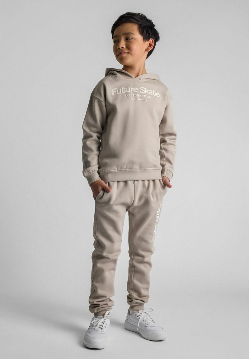 Beige sweatshirt and joggers set featuring "Future Skate" text, made of soft fabric, with ribbed cuffs and white sneakers.