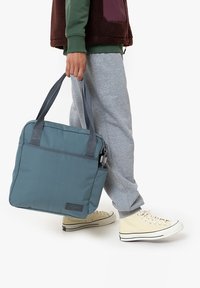 Borsa a tracolla grigia e blu realizzata in tessuto resistente, con due manici superiori e una tasca frontale con zip, portata da una persona in abbigliamento casual.