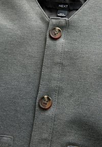 Tejido gris con una textura suave, presenta un collar redondeado, un cierre de botones con un diseño marmoleado y detalles cosidos.