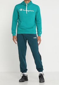Felpa con cappuccio color teal con logo "Champion" bianco, abbinata a pantaloni jogger teal scuro con dettagli neri e scarpe sportive nere.