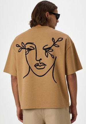 Homme portant un t-shirt oversize beige avec un dessin noir abstrait au trait d'un visage et des branches feuillues au-dessus des yeux dans le dos.