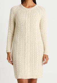 Krémové šaty s dlouhými rukávy a kulatým výstřihem vyrobené z pleteného svetru. Mají vroubkovaný lem a texturované vzory po celé tkanině.