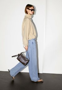 Beżowa koszula z długim rękawem, jasnoniebieskie szerokie dżinsy i czarne płaskie sandały. Trzyma małą, ciemnobrązową skórzaną torebkę z paskiem.