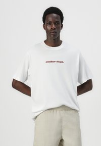 Camiseta de algodón blanca con cuello redondo y mangas cortas, con el texto rojo "atelier dept." impreso en la parte frontal.