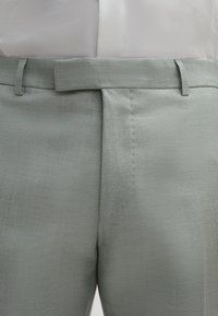 Hellgraue strukturierte Hose mit flachem Bund, schmaler Passform und Gürtelschlaufen, die dezente Nähtdetails entlang der Nähte aufweist.