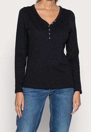 Frau trägt ein schwarzes Langarm-V-Ausschnitt-Shirt mit fünf Knöpfen und blaue Jeans, steht mit entspannten Armen an den Seiten.