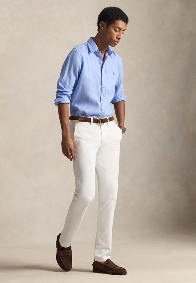 Ανοιχτό μπλε λινό πουκάμισο, λευκό παντελόνι, καφέ δερμάτινη ζώνη, καφέ σουέντ loafers. Το μοντέλο στέκεται σε ουδέτερο φόντο, κοιτάζοντας προς τα κάτω.