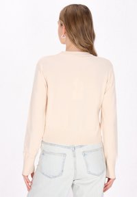 Šviesiai beige megztinis su raukšlėta apykakle ir rankogaliais, rodomas iš nugaros, derinamas su šviesiai mėlynomis džinsomis su kišenėmis.