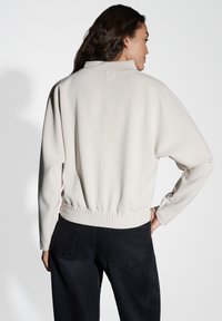 Sweatshirt cropped di un beige chiaro con colletto alto, vita a coste e spalle scese, abbinato a jeans scuri. Tessuto morbido.
