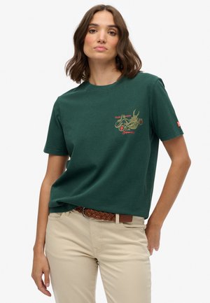 T-shirt en coton vert foncé à manches courtes, avec un design graphique doré et des accents rouges. Associé à un pantalon beige et une ceinture tissée marron.