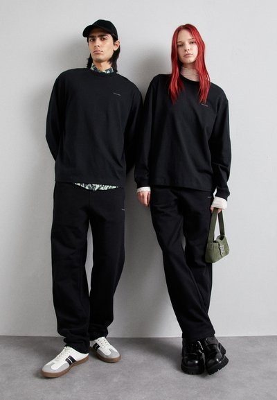 To modeller har matchende svarte hettegensere og bukser. Den mannlige modellen har på seg en svart caps og hvite joggesko; den kvinnelige modellen holder en grønn veske.