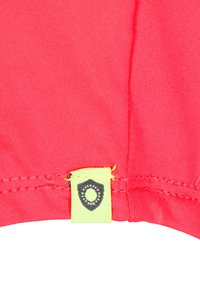 Tissu rose vif avec une texture côtelée. Présente une étiquette jaune avec un emblème foncé cousu à la couture, mettant en avant un détail de marque.