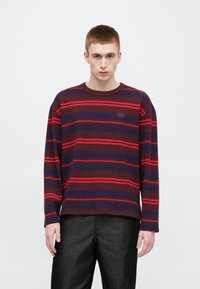 Μπλε σκούρο και μπορντό μακρυμάνικο ριγέ πλεκτό πουλόβερ με κόκκινες ρίγες, στρογγυλή λαιμόκοψη και επιγραφή με λογότυπο στο στήθος. Φορεμένο με σκούρο παντελόνι με υφή.